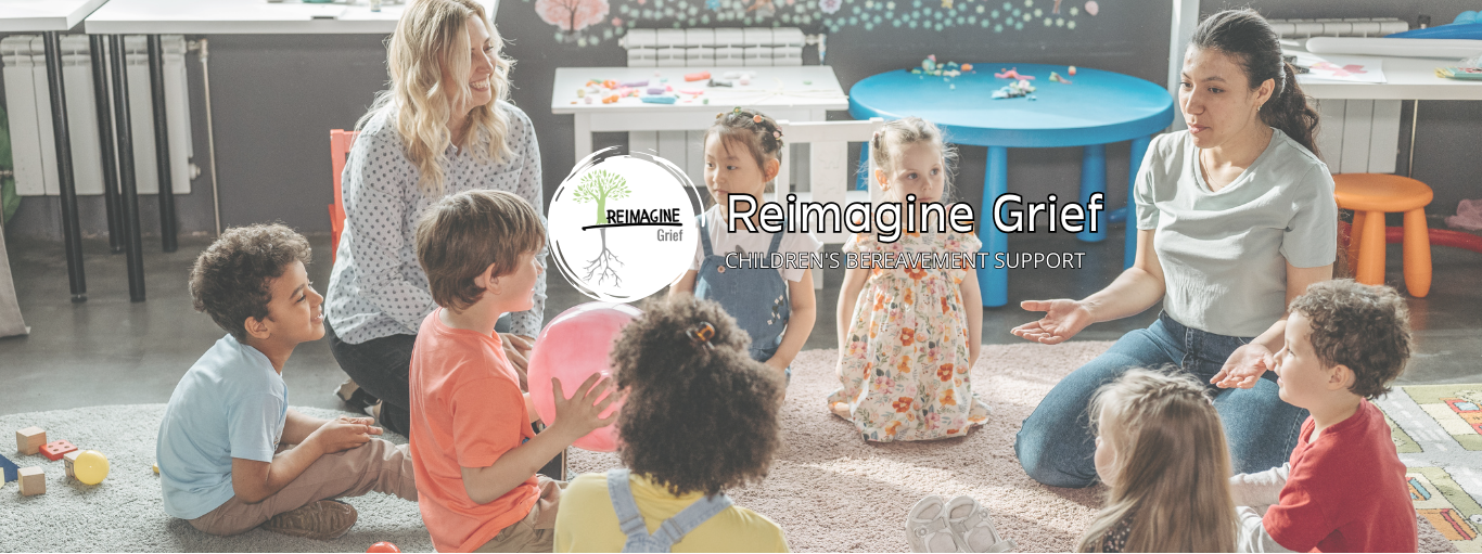 A group of children sit in a circle on the floor, smiling and playing a game together, while two adult teachers guide and encourage them.